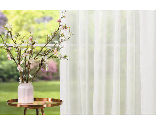 Crengi decorative cu flori într-o vază pe o măsuță laterală în fața unei ferestre cu draperie.