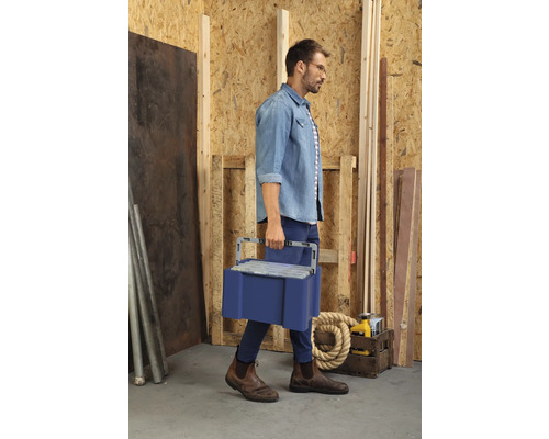 Un bărbat duce o cutie de scule albastră printr-un atelier