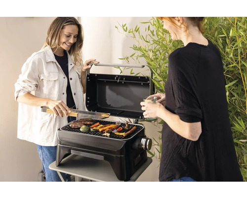 Două femei fac grătar cu legume și carne pe un grătar portabil în aer liber.