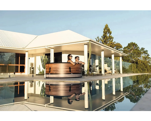 Scenă cu o cadă de baie din lemn lângă o piscină, în care se reflectă persoane și clădirea din jur.