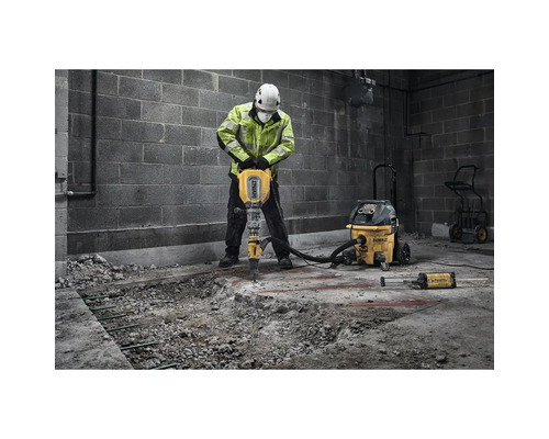 Un bărbat cu echipament de protecție folosește un ciocan pneumatic pentru a sparge beton. Un aspirator și un cărucior sunt în apropiere.