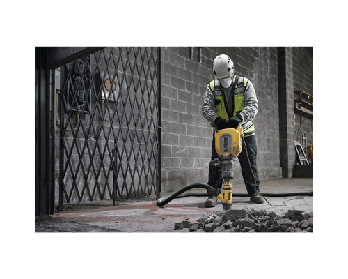 Un muncitor cu cască de protecție și vestă de siguranță operează un ciocan pneumatic galben pe o podea de beton.