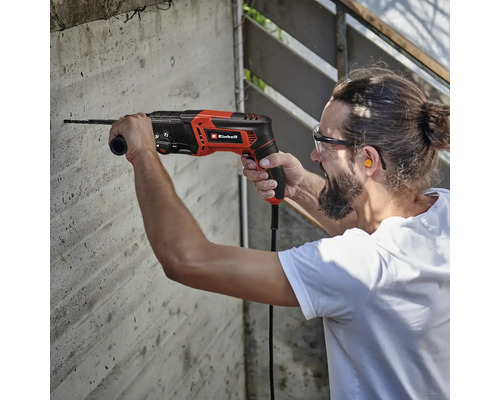 Un bărbat forează o gaură într-un perete de beton cu un ciocan rotopercutor, sigla Einhell.