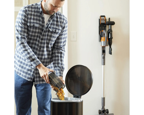 Un bărbat golește recipientul de praf al unui aspirator fără fir într-un coș de gunoi. Pe perete este o stație de încărcare cu un aspirator.