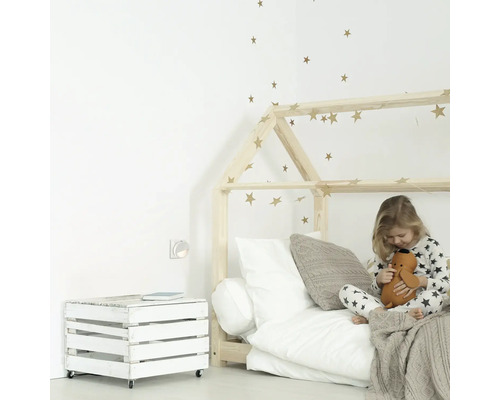 Cameră luminoasă pentru copii cu pat din lemn în formă de casă, ladă din lemn lăcuită alb pe roți și o fetiță care se joacă cu o jucărie de pluș