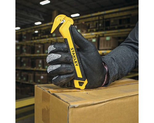 O mână cu o mănușă de protecție ține un deschizător de scrisori Stanley pe o cutie de carton.