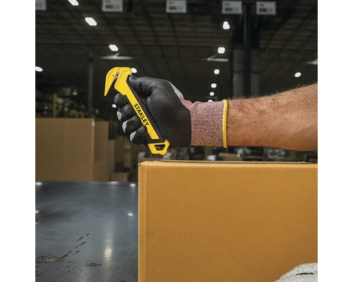 O mână cu mănușă folosește un tăietor de carton Stanley galben pentru a deschide o cutie de carton.
