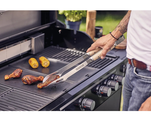 Un bărbat prăjește pulpe de pui și știuleți de porumb cu un clește de grătar pe un grătar