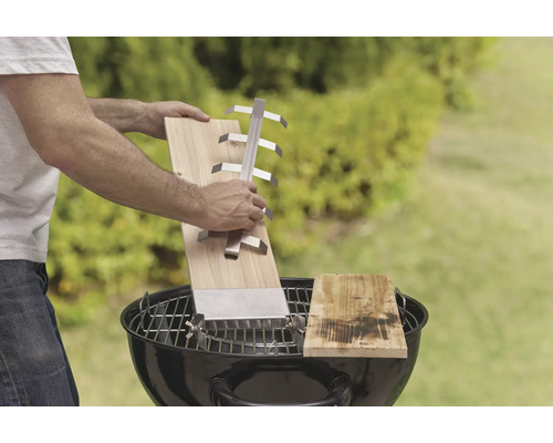 Scenă cu un bărbat ținând o scândură de grătar cu suport deasupra unui grătar cu cărbune, în aer liber.