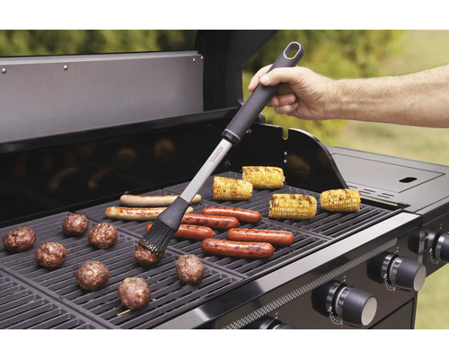 Scenă de grătar cu perie de grătar, cârnați, știuleți de porumb și chiftele pe un grătar pe gaz.