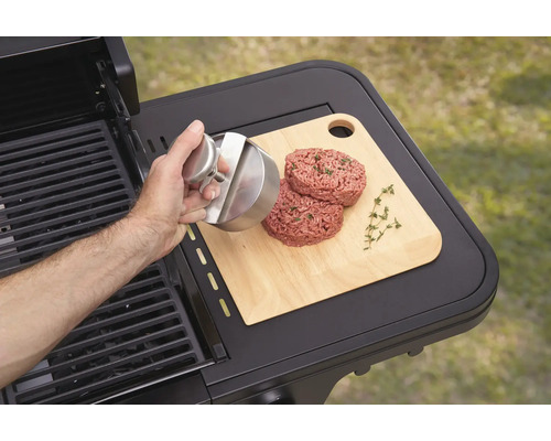 O persoană formează burgeri cu o presă de burgeri pe un tocător lângă un grătar.