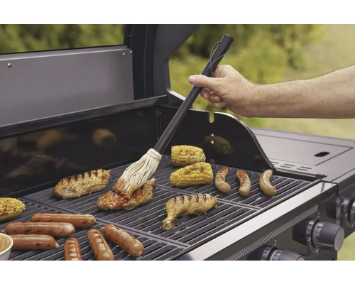 Un bărbat unge carne cu un sos pe un grătar cu știuleți de porumb și cârnați.