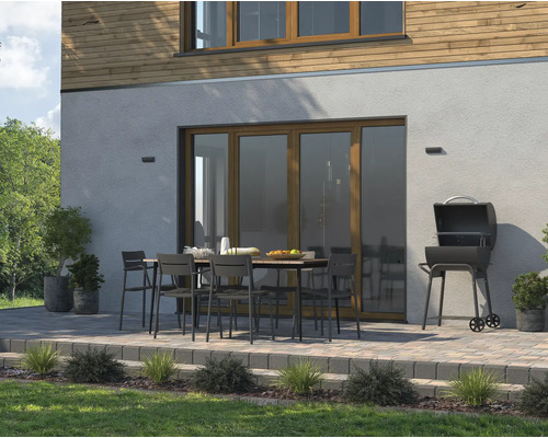 Scenă exterioară cu grătar, masă de grădină și scaune pe o terasă în fața unei case