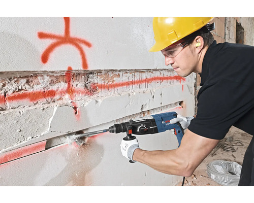 Un bărbat găurește un perete cu un ciocan rotopercutor. Poartă echipament de protecție.