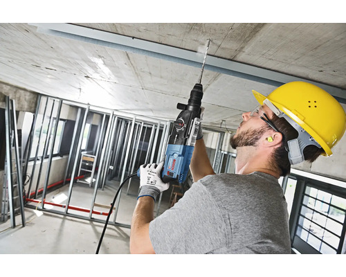 Un bărbat forează cu o mașină de găurit într-un tavan de beton, poartă o cască de protecție.