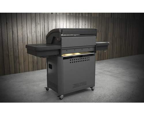 Grătar pe roți gri închis, cu rafturi laterale și role în fața unui perete de lemn