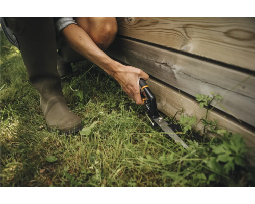 O persoană care folosește foarfece de tuns iarba pentru a tăia iarba de lângă un perete de lemn