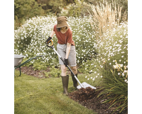 Femeie lucrează cu o sapă în grădină, înconjurată de flori și verdeață.