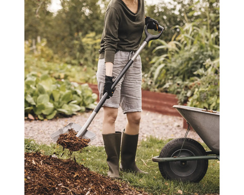 Femeie care folosește lopata pentru a muta mulci într-o grădină.
