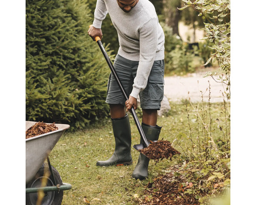 O persoană manevrează o lopată în grădină și încarcă mulci într-un cărucior.