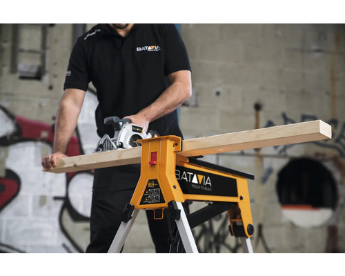 Artizan care taie o scândură de lemn cu un ferăstrău circular manual pe un banc de lucru. Logo Batavia.