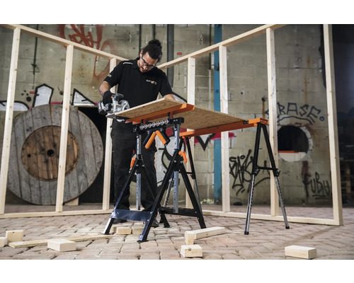 Un bărbat taie lemne pe un banc de lucru cu un ferăstrău circular portabil. În fundal sunt grinzi de lemn și un tambur de lemn.