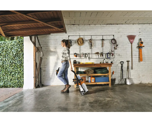 Femeie împinge un aparat de spălat cu presiune pe roți într-un garaj cu unelte de grădină.