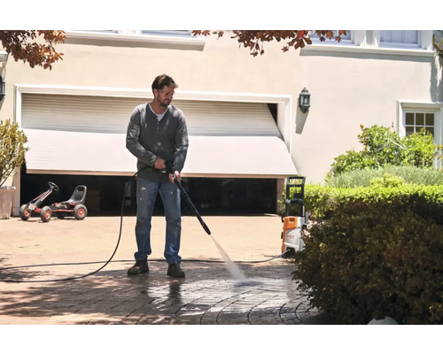 Bărbat curăță pavajul cu un curățător cu presiune în fața unui garaj.