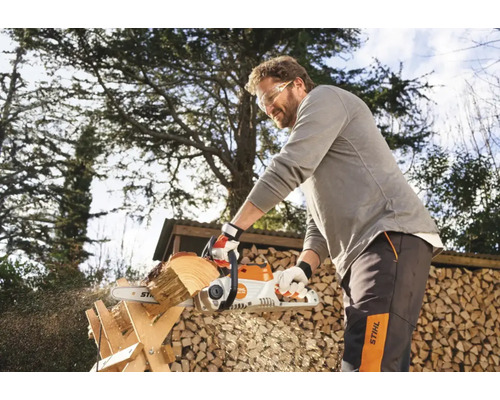 Un bărbat taie lemne cu un motoferăstrău afară, în fața unui depozit de lemne.