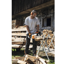 Un bărbat taie lemne cu un lanț de ferăstrău pe un suport de tăiere, purtând echipament de protecție, cum ar fi protecție pentru urechi, ochelari de protecție și mănuși.