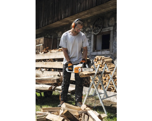 Un bărbat taie lemne cu un lanț de ferăstrău pe un suport de tăiere, purtând echipament de protecție, cum ar fi protecție pentru urechi, ochelari de protecție și mănuși.