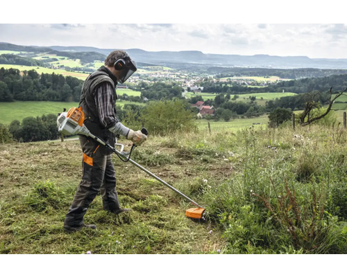 Un bărbat lucrează cu un motocoasă pe un câmp. În fundal se vede un peisaj deluros.