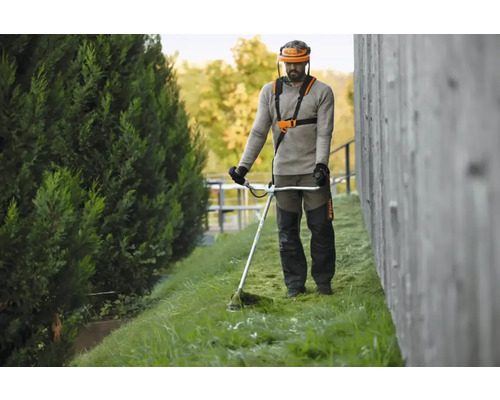 Bărbat operează motocoasă cu vizieră de protecție și curea de transport în grădină