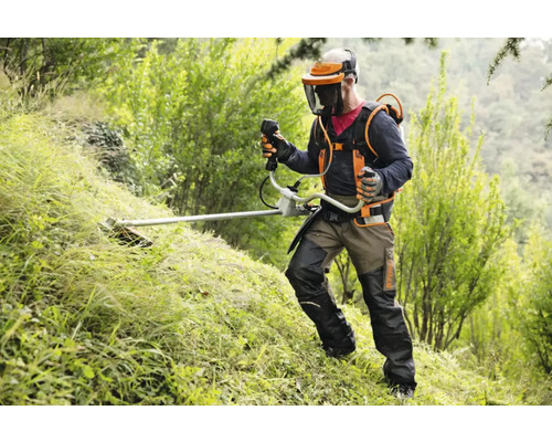 Bărbat cu cască de protecție și motocoasă grădinărind în verdeață.