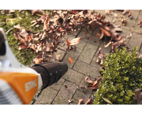 Suflantă de frunze în funcțiune pe o suprafață pavată cu frunze împrăștiate.