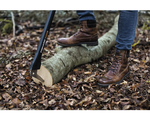 Scenă cu un topor Fiskars înfipt într-un trunchi de copac și o persoană cu cizme stând pe trunchi.