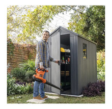 Scenă de grădină cu un bărbat ținând o suflantă de frunze și deschizând o magazie de grădină cu rafturi și unelte de grădină.