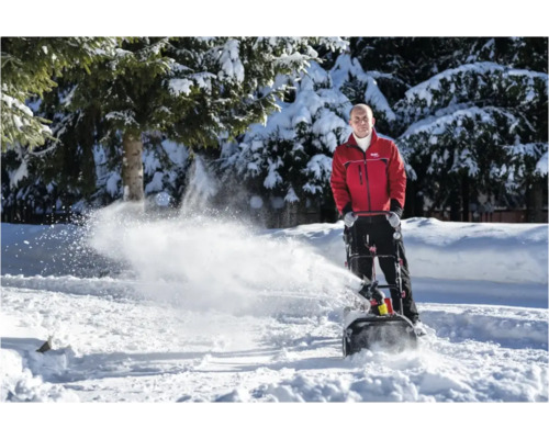 Un bărbat manevrează o freză de zăpadă pe o suprafață acoperită de zăpadă, cu copaci în fundal.