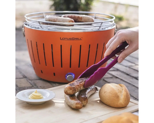 Grătar LotusGrill cu alimente la grătar, clește, chiflă și sos pe o masă de lemn