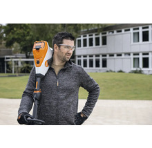 Un bărbat cu ochelari de protecție poartă un trimmer de gazon cu acumulator Stihl pe umăr.
