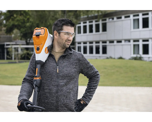 Un bărbat cu ochelari de protecție poartă un trimmer de gazon cu acumulator Stihl pe umăr.