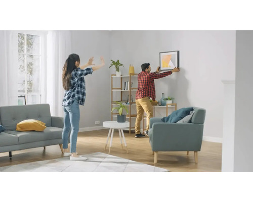 Living luminos cu un cuplu care agață un tablou pe perete, rafturi și mobilier de ședere