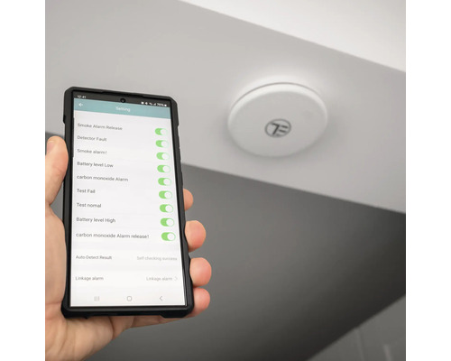 Un detector de fum este montat pe tavan în timp ce un smartphone cu un afișaj de setări este ținut în mână