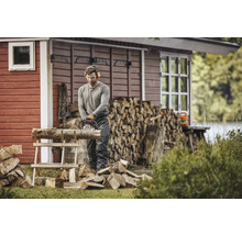 Un bărbat taie lemne cu un motoferăstrău în fața unei case de lemn