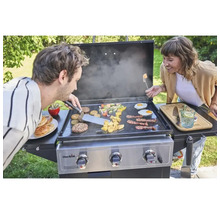 Două persoane pregătesc mâncare pe o plită Char-Broil, inclusiv legume, burgeri, ouă și bacon.