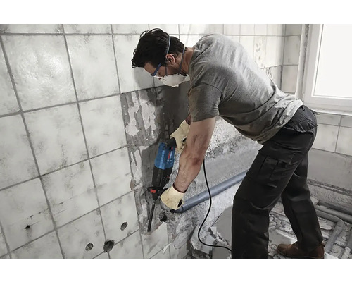 Un bărbat lucrează cu un ciocan rotopercutor pe un perete placat cu faianță într-o baie.