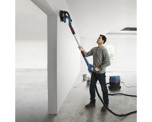 Un bărbat șlefuiește un perete cu o șlefuitoare cu gât lung într-o cameră. Un aspirator se află în fundal.