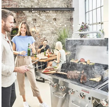 Scenă cu un grătar, cu un grătar din oțel inoxidabil folosit pentru a prepara carne și legume, în timp ce prietenii și familia stau la o masă în fundal.