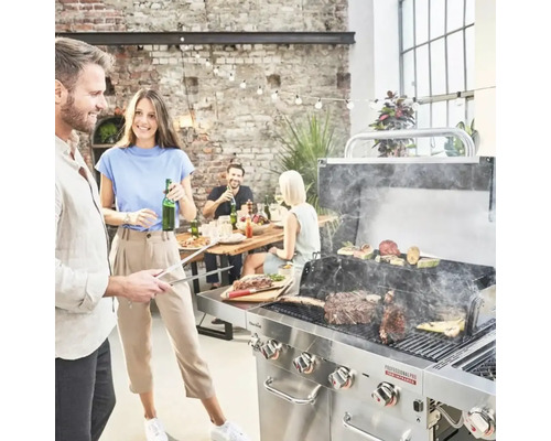 Scenă cu un grătar, cu un grătar din oțel inoxidabil folosit pentru a prepara carne și legume, în timp ce prietenii și familia stau la o masă în fundal.