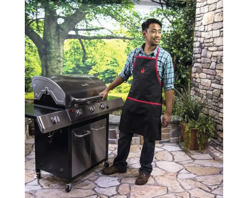 Bărbat cu șorț de grătar stând lângă un grătar pe gaz în grădină.